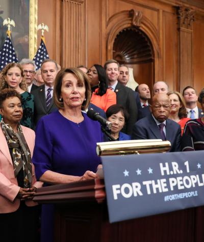 Nancy Pelosi standing a a podium on which is written the words "H.R. 1 For the People Act" with a large group of people standing around and behind her.